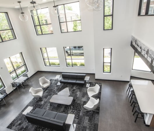Overhead view of indoor community space with black couches, white chairs, two-person tables and a counter space with black barstools. The two-floor space has windows from top to bottom letting in a lot of natural light.