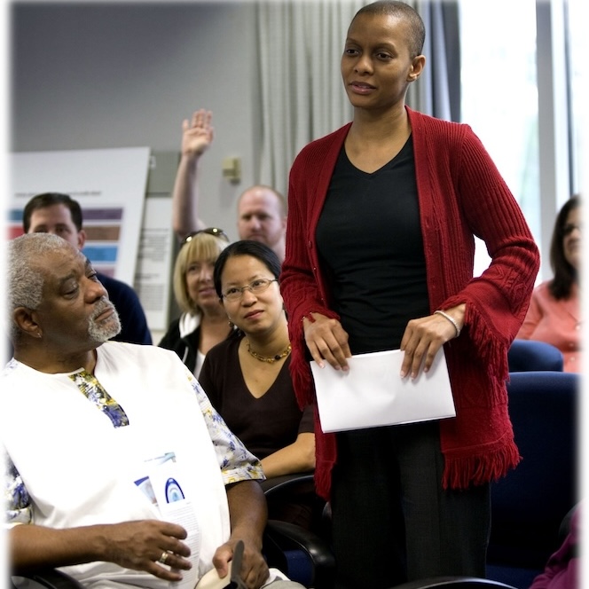 A woman standing in a crowd of people holding a white paper and talking