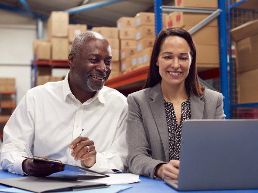 A middle-aged African American businessman and middle-aged business woman look at a laptop screen together. They are sitting in a warehouse with boxes stacked on shelves behind them.
