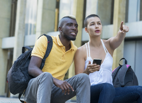 A young African American man and a young white woman sit together on some
steps with their backpacks. The woman is holding a phone and pointing to something off in the distance.