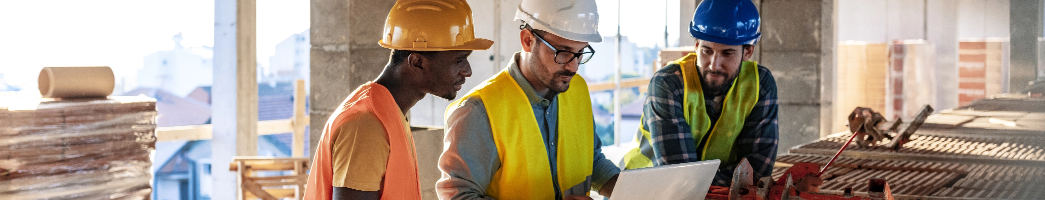Three working men in hard hats and bright yellow vests stand in a building looking at
a laptop together.