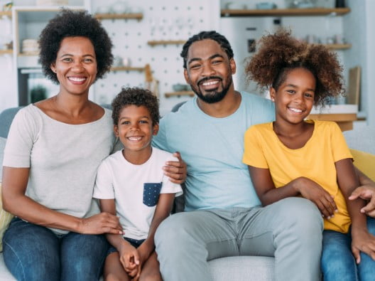 An African American family of four sits smiling on their couch in their home. It
consists of a woman, man, young girl and a slightly younger boy.